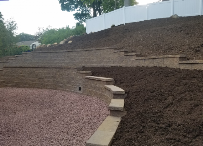 A terraced landscape with stone walls, stairs, and dark soil. A white fence is in the background.