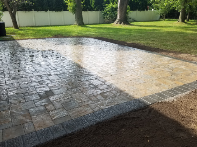 Patio with stone pavers and edging, partially wet, in a grassy backyard, surrounded by trees and a white fence.
