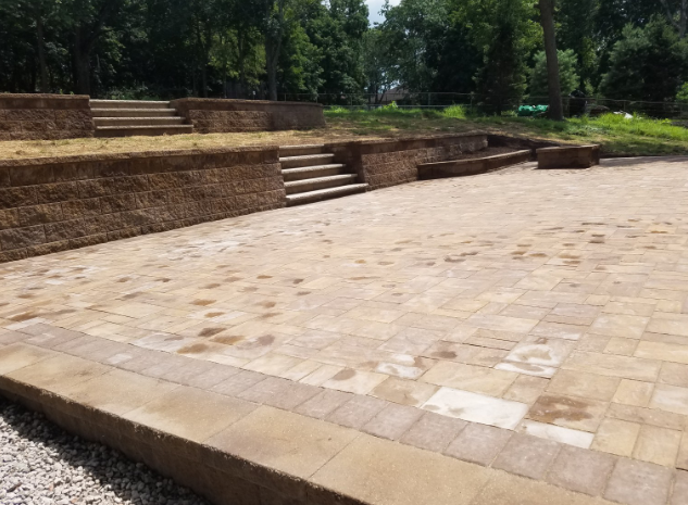 Outdoor paved patio with terraced seating and retaining walls, leading to trees.