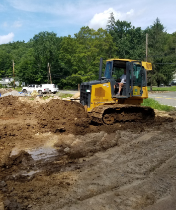 Yellow bulldozer on muddy ground, pushing dirt. Green trees in background, sunny day.