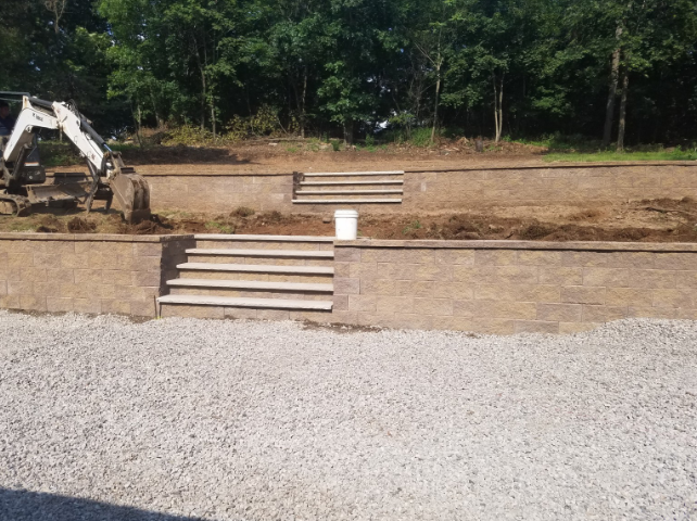 Stone retaining walls with steps, gravel driveway, and a small excavator, next to a wooded area.