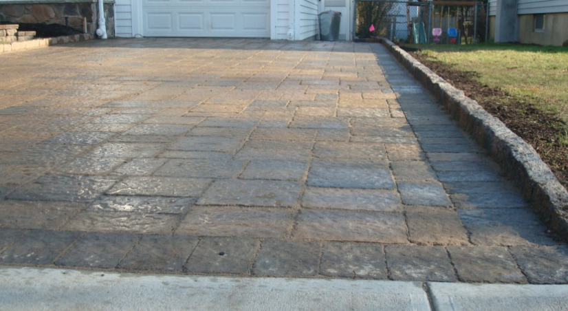 Stone paver driveway leading to a white garage, bordered by a concrete curb and landscaping.