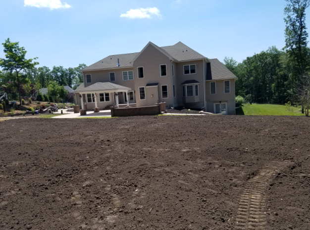 Back of a two-story beige house with a large yard freshly tilled, under a bright blue sky.
