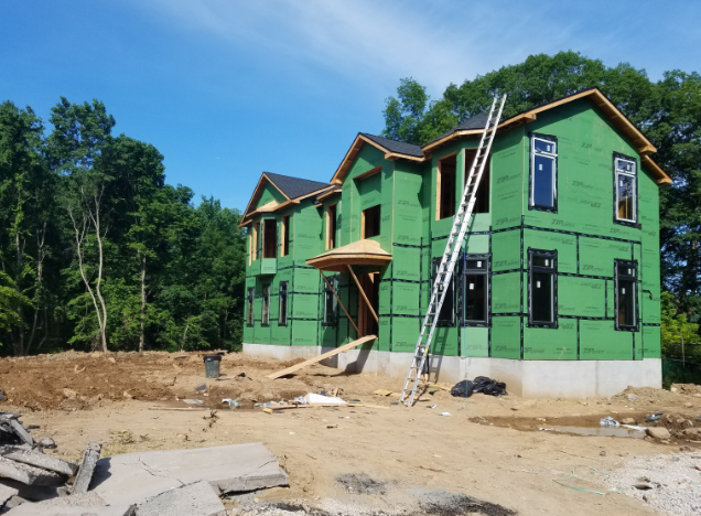 Two-story house under construction, green sheathing, dark windows, ladder, sunny day.