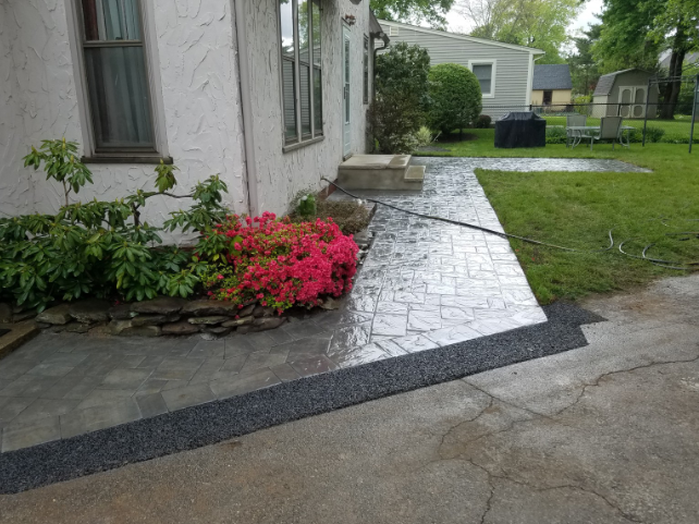 Concrete walkway with pink flowers next to a house. Dark gravel borders the path.