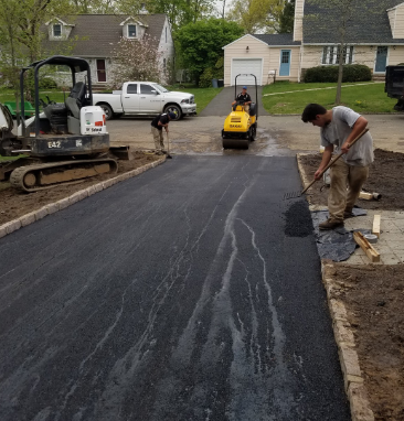 Asphalt driveway paving in progress. Workers spread and compact blacktop. Machinery and houses in background.