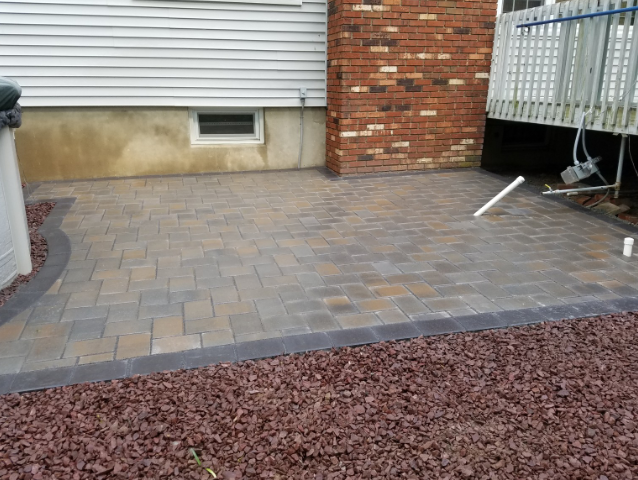 Brick patio with dark border, next to house with brick chimney and gravel.