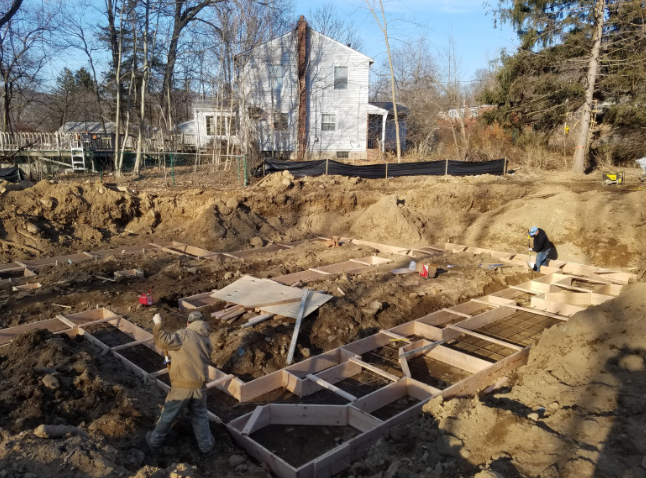 Construction site with wooden forms for foundation. Two workers, one inside the forms. Earth, house, and trees in background.