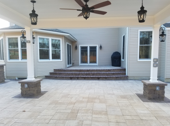 Covered patio with a paver floor, steps, and house with windows. Lanterns and a ceiling fan are present.
