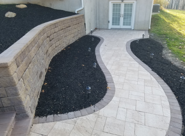 Stone pathway curves through black mulch, beside a retaining wall and building with glass doors.