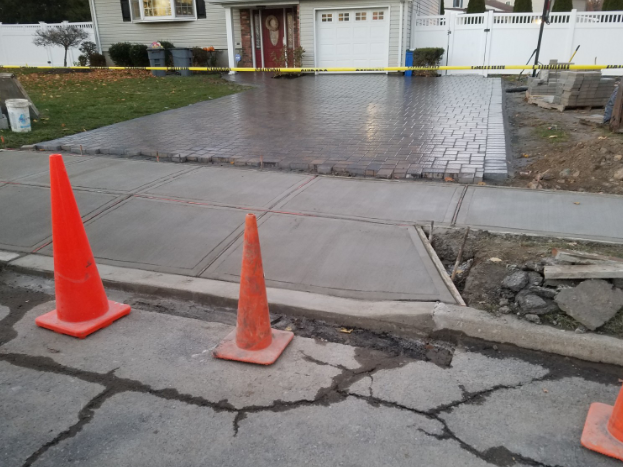 Orange cones on a cracked sidewalk next to a newly paved driveway and a house with a white garage door.