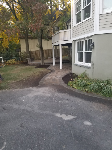 Asphalt driveway leads to a gray stone pathway winding toward a house with a deck.