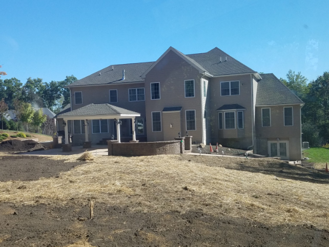 Back view of a large two-story house with a covered patio, set on a slightly sloped yard, under a blue sky.