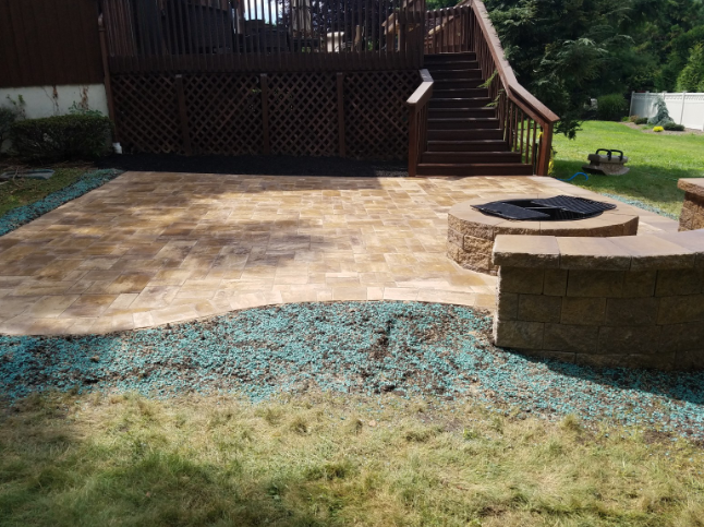 Stone patio with built-in fire pit, connected to a wooden deck with stairs. Lawn area in foreground.