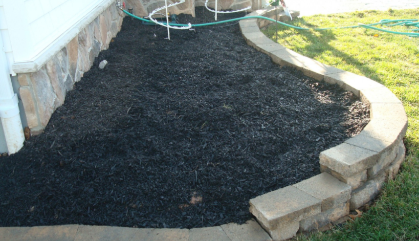 Black mulch covers a stone-edged garden bed against a white building and grass lawn.