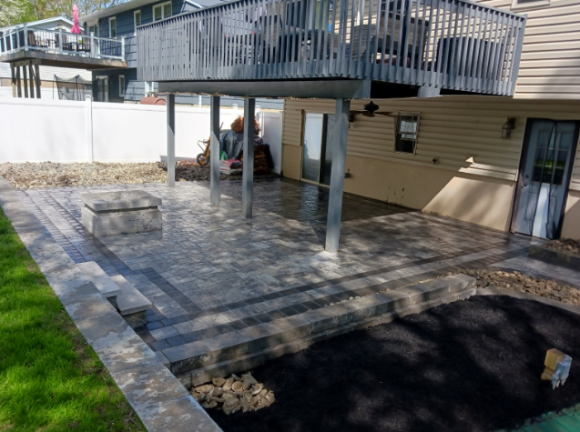 Paver patio under a deck. Gray pavers, black border. Overlooking a yard with green grass.