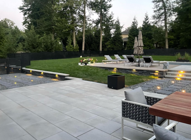 Patio with pool, lawn, and trees. Gray stone, lounge chairs, and outdoor lighting.
