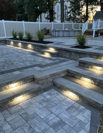 Stone patio with built-in steps and lights. Landscaping with shrubs in a backyard setting.
