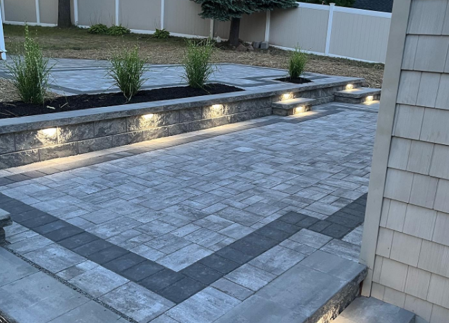 Brick patio with retaining wall, built-in lights, and planted greenery. Dark gray pavers and tan siding.
