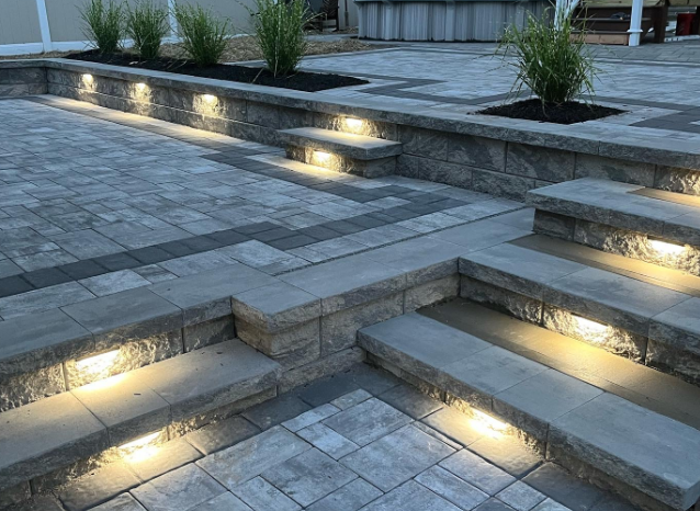 Stone patio with built-in steps and retaining walls, illuminated by warm lighting, with plants in raised beds.