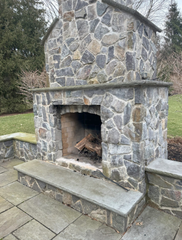 Stone outdoor fireplace with a raised hearth, logs inside, set on a stone patio.