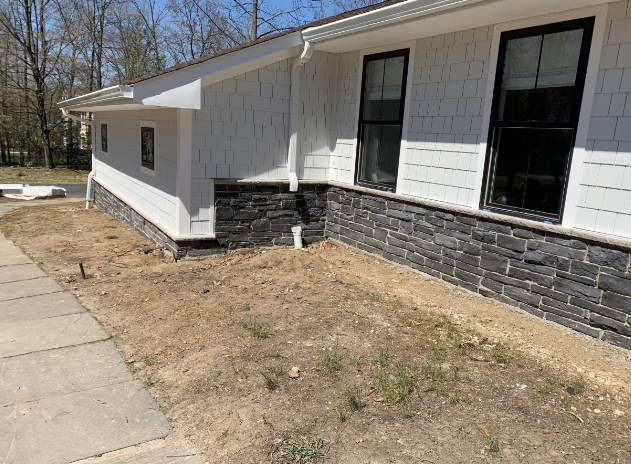 Exterior view of a house with white siding and black stone veneer on the bottom. Brown dirt yard in front.