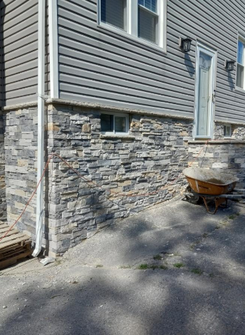 Gray house with stone veneer base and gray siding, white trim, and a small basement window.