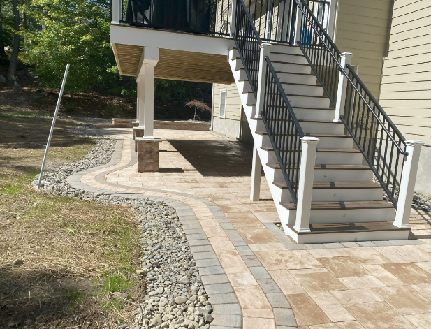 Paved patio with steps leading up to a deck, bordered by a rock garden. The house has tan siding.