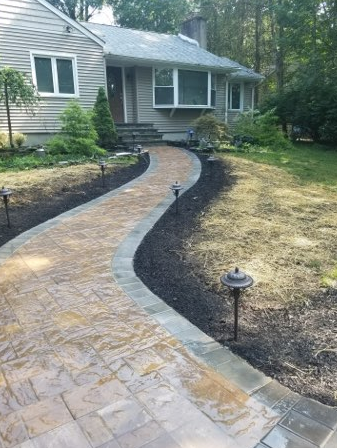 Brick walkway leading to a gray house, lined with landscaping and low lights.
