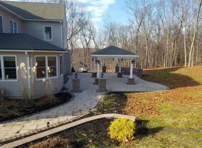A paved patio with a gazebo, connected by a walkway to a house on a sunny day.