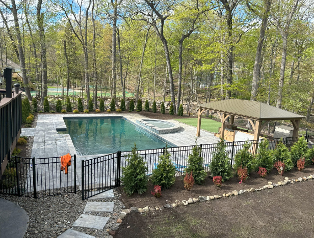 Backyard pool area with a dark-tiled pool, a hot tub, and a wooden gazebo. Fencing, trees, and landscaping surround the area.