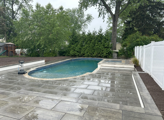 Pool and patio area with blue water, gray pavers, white fence, and greenery.