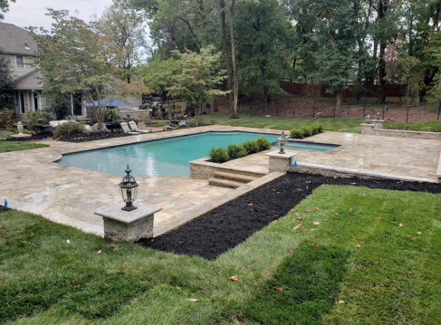 Backyard pool with stone patio, lawn, and dark mulch beds; trees in the background.