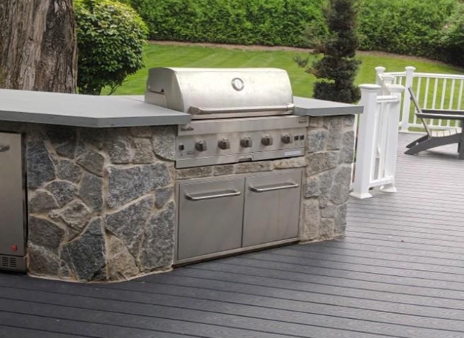 Outdoor kitchen with stainless steel grill, stone facade, and gray countertop on a deck.