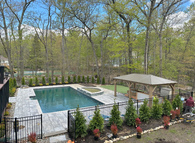 Backyard pool with a gazebo, landscaping, and a black fence. Trees in the background.