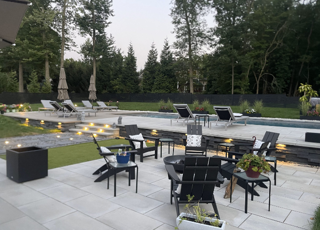 Outdoor patio with seating area by a pool, surrounded by greenery and trees.