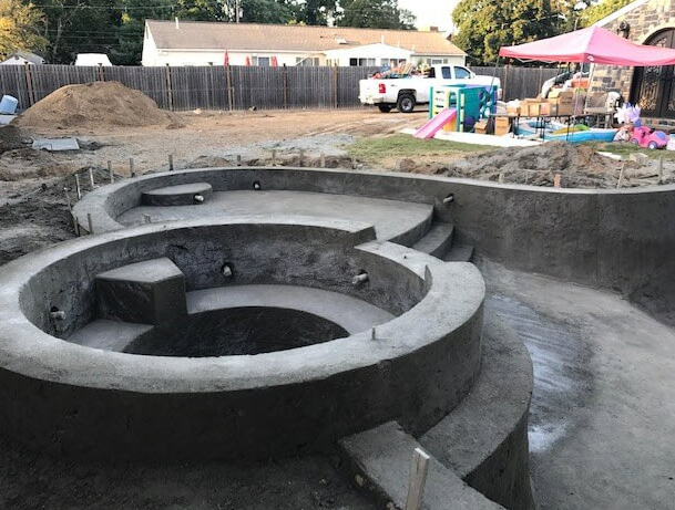 Pool construction site, concrete pool shell with circular features and steps, outdoor setting.