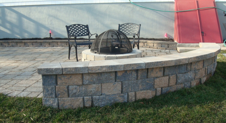 A curved, brick patio with a fire pit, two chairs, and a retaining wall.