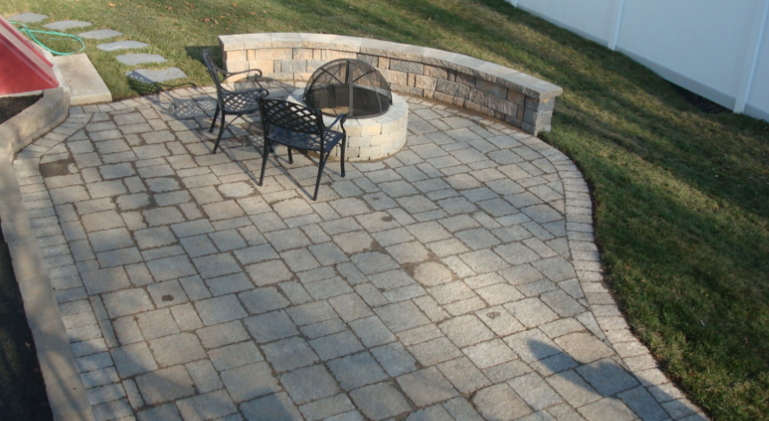 Brick patio with fire pit, seating, and a retaining wall. Green grass surrounds.