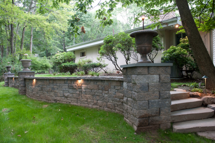 Stone wall with urns, built-in lights, and steps leading to a house entrance, surrounded by greenery.