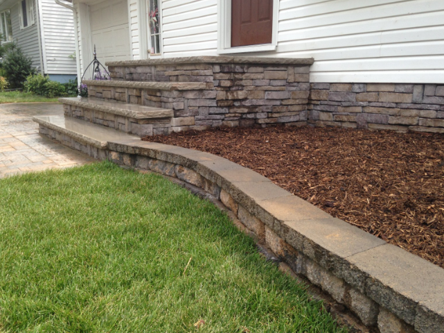 Stone steps and retaining walls in front of a white house with a brown door. Green grass and mulch.