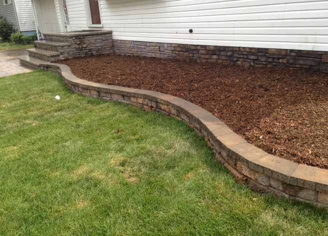 Stone retaining wall with a mulch bed, in front of a white house. Green lawn surrounds the wall.