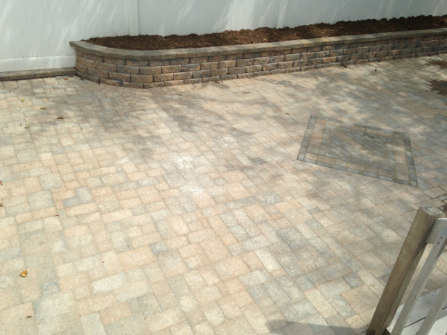 Brick paver patio with a low retaining wall holding soil, against a white fence.