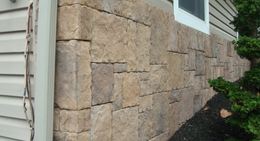 Stone veneer siding on a building's foundation, tan and brown shades, next to a window and a bush.