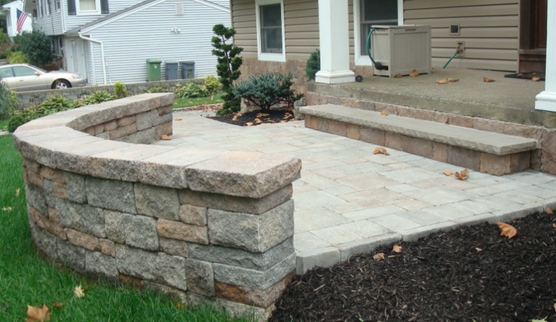 Stone patio with curved retaining wall and bench next to a house with steps.