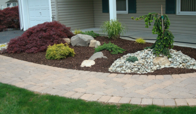 Landscaped front yard with brick pathway, mulch, rocks, and various plants.