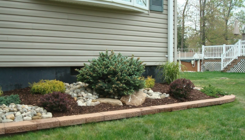 A landscaped garden bed with mulch, rocks, and various plants borders a house with tan siding and a white deck.