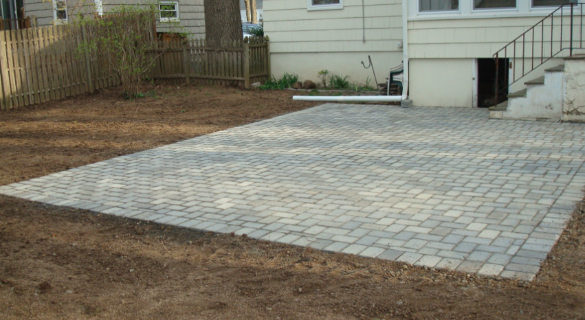 A gray brick patio in a backyard setting next to a beige house, steps, and a brown fence.