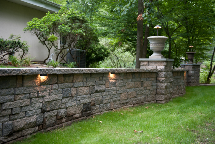 Stone retaining wall with built-in lights, topped with urns and greenery, along a grassy lawn.