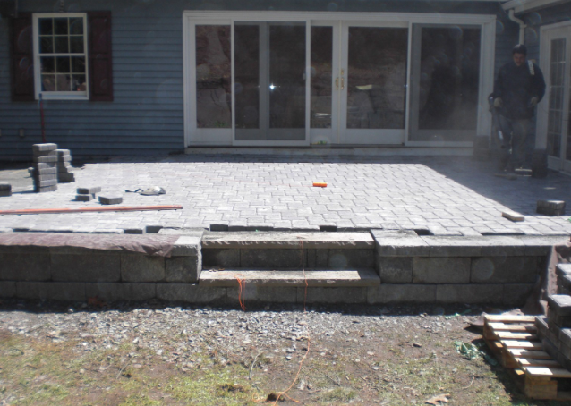 Patio construction: Brick pavers, retaining wall with steps, next to a house with a sliding glass door.